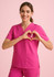 A woman in a bright pink V-neck scrub top makes a heart shape with her hands. The top has a logo.