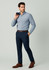 A man in a light blue shirt and navy slim fit flat front pants, holding a dark blue item, wearing brown shoes.