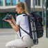 A woman with a black Swissdigital Berg XL Backpack crouches, holding a camera. The backpack has a tripod attached.