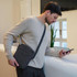 A shoulder bag in grey with a shoulder strap, held by a man in a light sweater, looking at his phone.