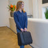 A woman holds a black wheeled underseat luggage bag in a stylish indoor setting. The bag has a logo.