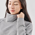 A woman wearing a grey cowl neck pullover, featuring long sleeves and a relaxed fit. The pullover has a logo.