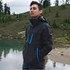 A man stands by a body of water, wearing a black softshell jacket with blue zippers in a natural setting.