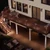A two-story house features a long elevated wooden deck extending along the back of the home. Warm lighting highlights the deck railing, stairs, and lower patio seating area at dusk. Multiple windows reveal the interior glow, while outdoor furniture and potted plants create a welcoming gathering space. The surrounding landscaping frames the deck and enhances the cozy evening atmosphere.
