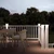 An outdoor deck with white railings is softly illuminated at dusk, overlooking a dense line of trees. A round wooden table with woven chairs sits on the warm-toned deck surface, suggesting a cozy gathering space. Built-in post lights glow along the railing and stairs, enhancing safety and ambiance. The sky is fading into twilight, creating a calm and inviting atmosphere for outdoor relaxation.