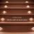 Close-up view of wooden deck stairs featuring small recessed step lights along each riser. The lights emit a soft white glow, evenly spaced to highlight the stair edges. Text in the center reads "Compatible by Design" and "Works with all deck stairs," emphasizing versatility. The overall scene is warm and modern, focusing on outdoor safety and style.
