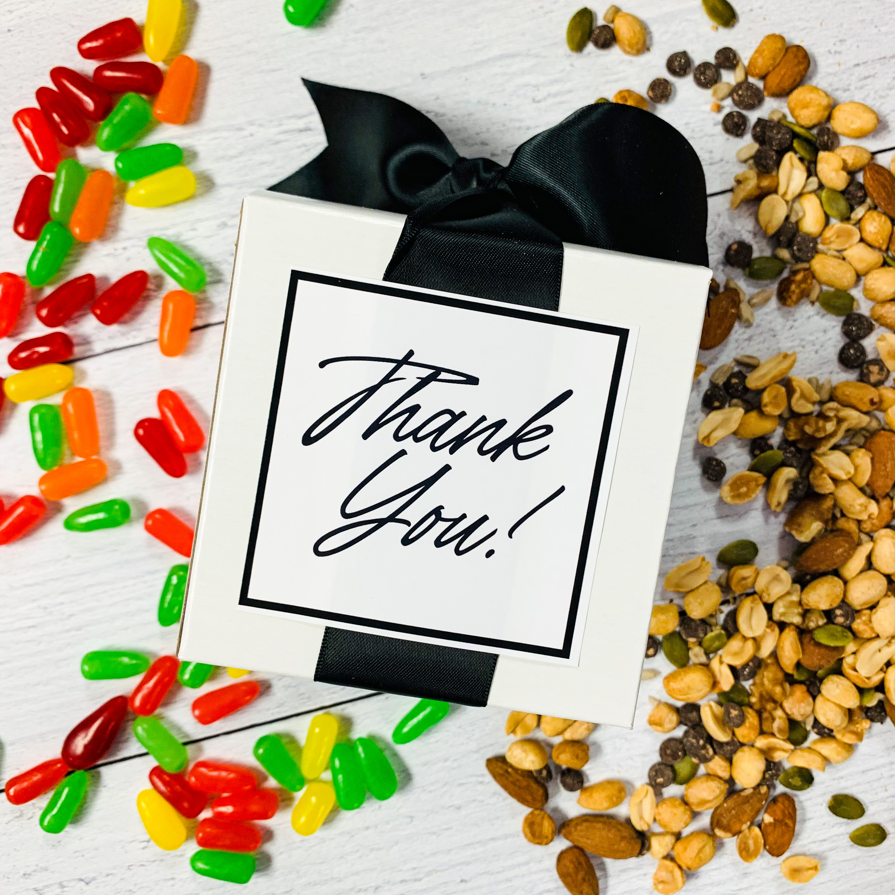 A white cube box with a black bow and "Thank You" on the top face surrounded by Mike and Ikes and granola