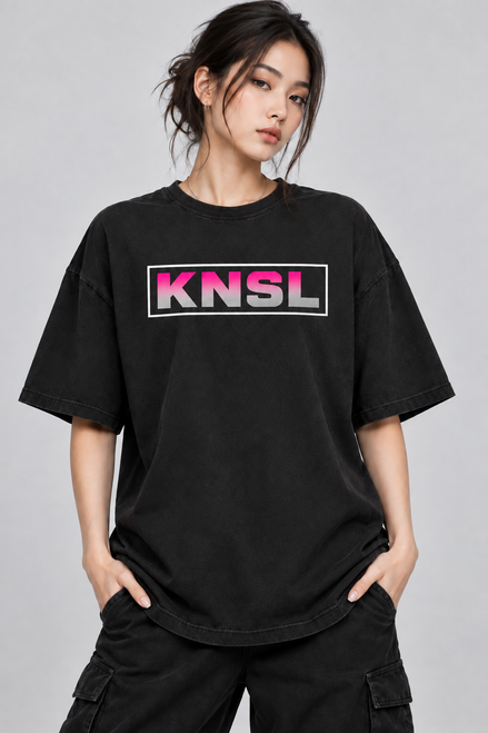 emale model wearing a black, regular-fit t-shirt. The chest features a graphic with the letters "KNSL" in a grey-to-hot-pink gradient inside a white rectangular border.