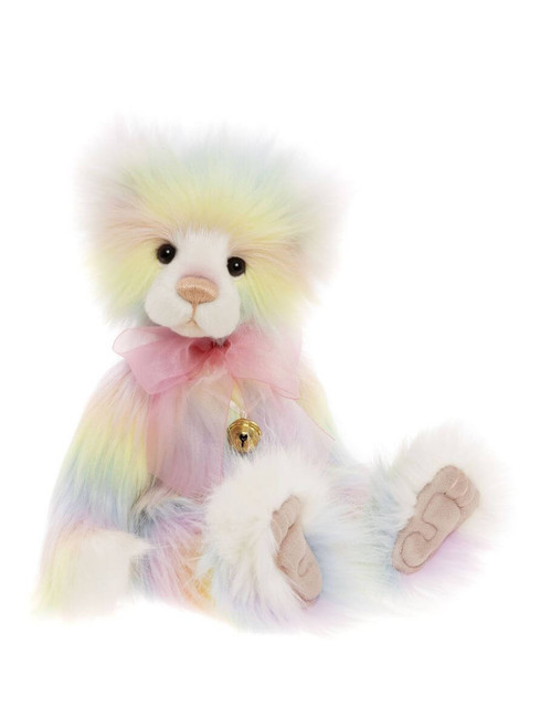 A pastel rainbow-coloured fluffy teddy bear sits upright against a white background, wearing a pink organza bow and small gold bell around its neck.