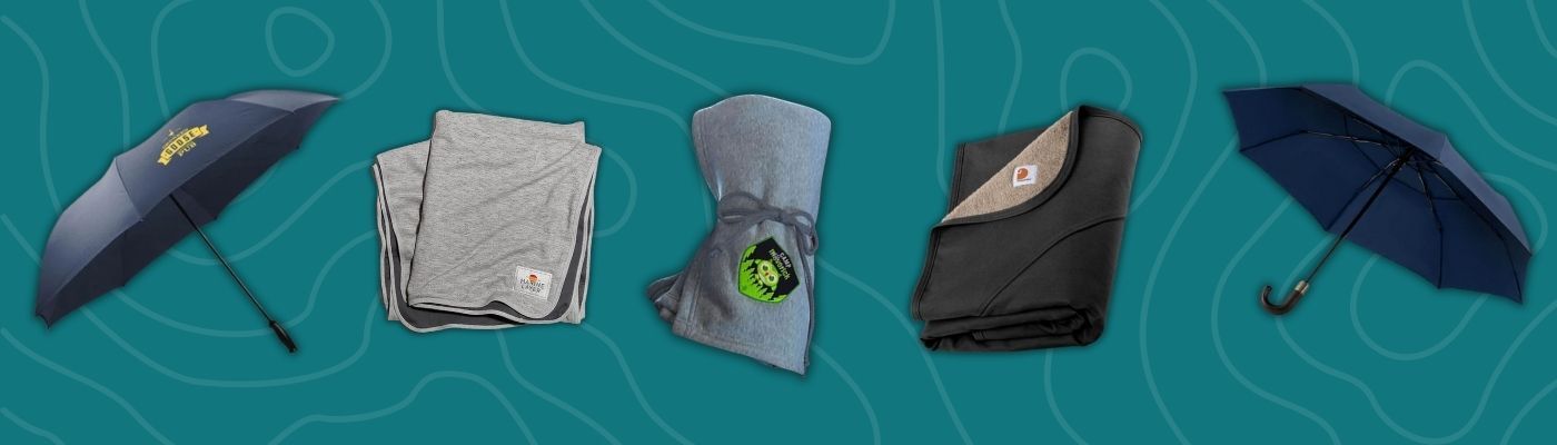 Assortment of ShedRain umbrellas, Carhartt and Marine Layer blankets, and Tumi lifestyle gear featuring the best custom promotional products for outdoor use.