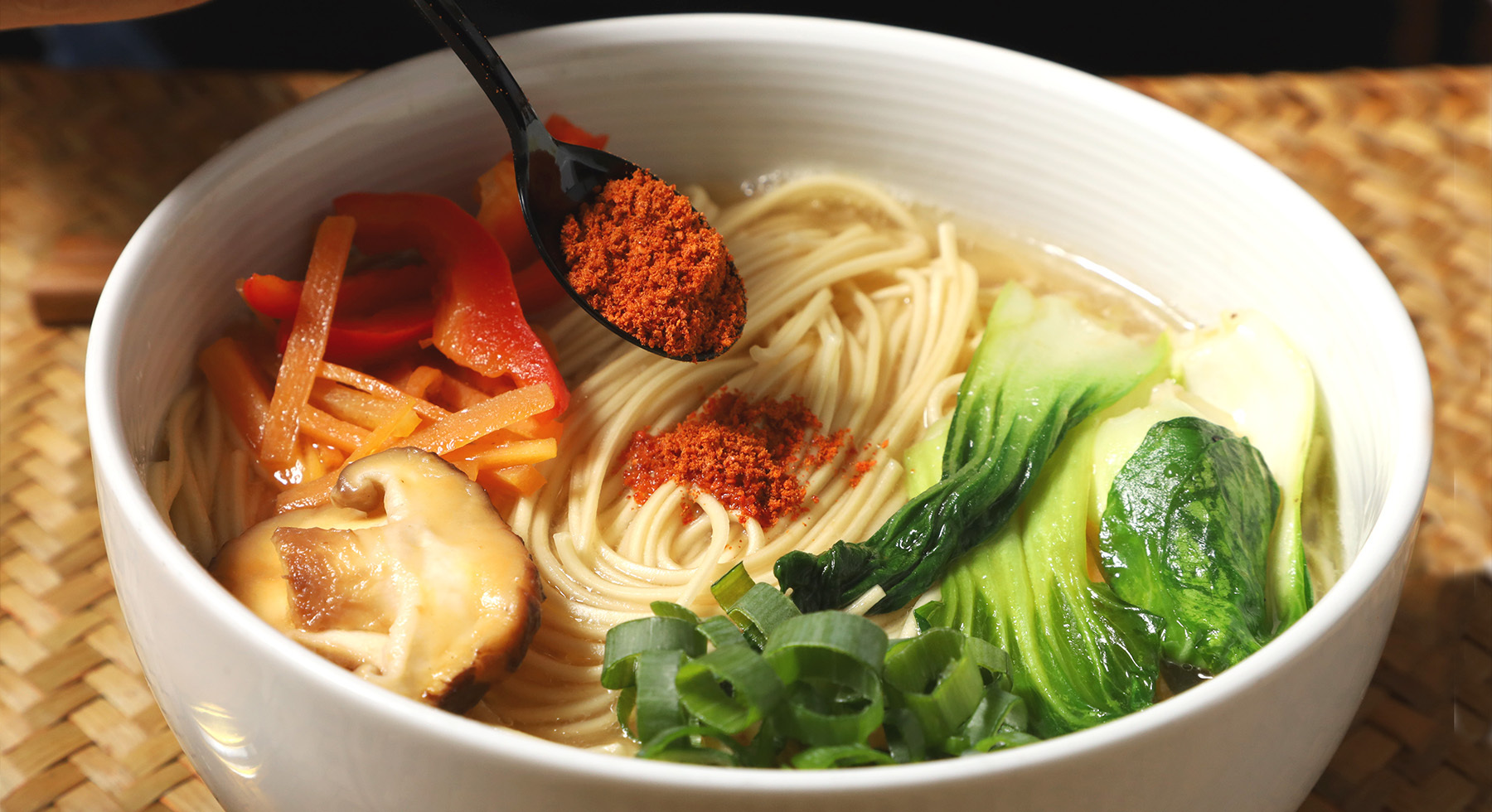 Close up of a small black spoon placing a generous amount of the  Original Spicy Red Seasoning powder onto a bowl of vegan ramen topped with bok choy, shiitake mushrooms, sliced carrots, and red bell peppers.