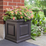 Presidential Square Planter, a large black decorative flower planter filled with pink and yellow flowering plants placed outside near a brick wall and garden.