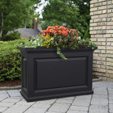 Presidential Trough Planter, black rectangular outdoor planter filled with colorful flowers on a paved patio near a brick wall and green trees.