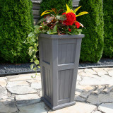 Promenade Tall Patio Planter, a tall gray rectangular outdoor planter with decorative paneling, filled with colorful flowers, placed on a stone patio with green bushes in the background.