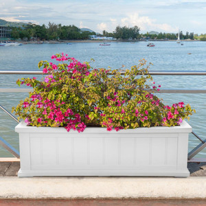 Promenade Railing Planter, white decorative flower box filled with vibrant pink and purple flowering plants, situated on a waterfront promenade with a view of water and boats in the background.