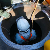 A worker with a white hard hat on going down a manhole with another worker in green looking on