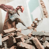 A worker in orange hard hat, wearing gloves using a jackhammer to demolish a brick wall