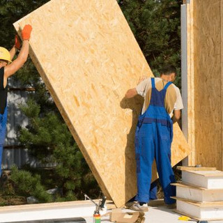 Two men, one wearing gloves and hardhat carrying a piece of wood panelling.