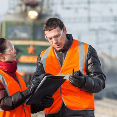 Man in high vis vest & safety glasses looking over a checklist with a train in the background.