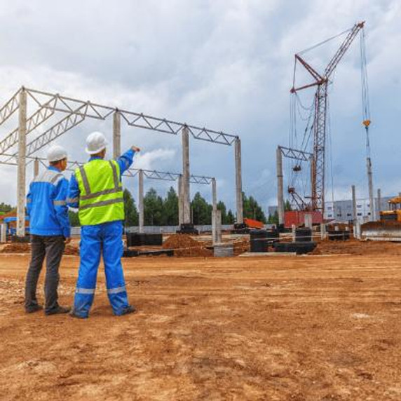 Two workers on a worksite, one in a blue jacket the other in a yellow high-vis, both wearing white hard hats discussing the construction job in front of them. One worker pointing off in the distance.