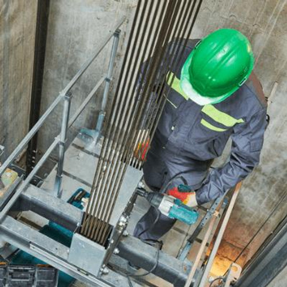Man in a lift shaft wearing a green hardhat and drilling the suspensions.