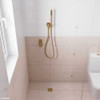 Urban brass square slim floor waste with matching mixer and shower head on a hook featured in a bathroom with light pink and white tiles.