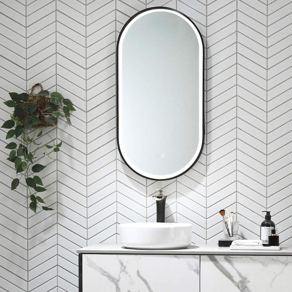 Black pill shaped LED mirror mounted above a bathroom vanity on a white herring bone tile.