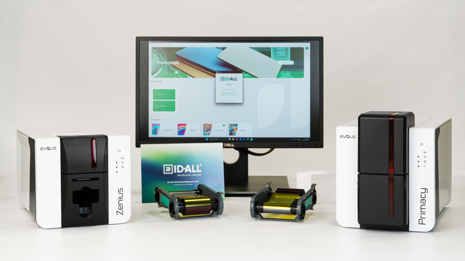 Evolis Primacy 2 card printer setup with a desktop monitor displaying card design software, two Primacy 2 ID card printers on either side, and stacks of printed plastic ID cards arranged in front.