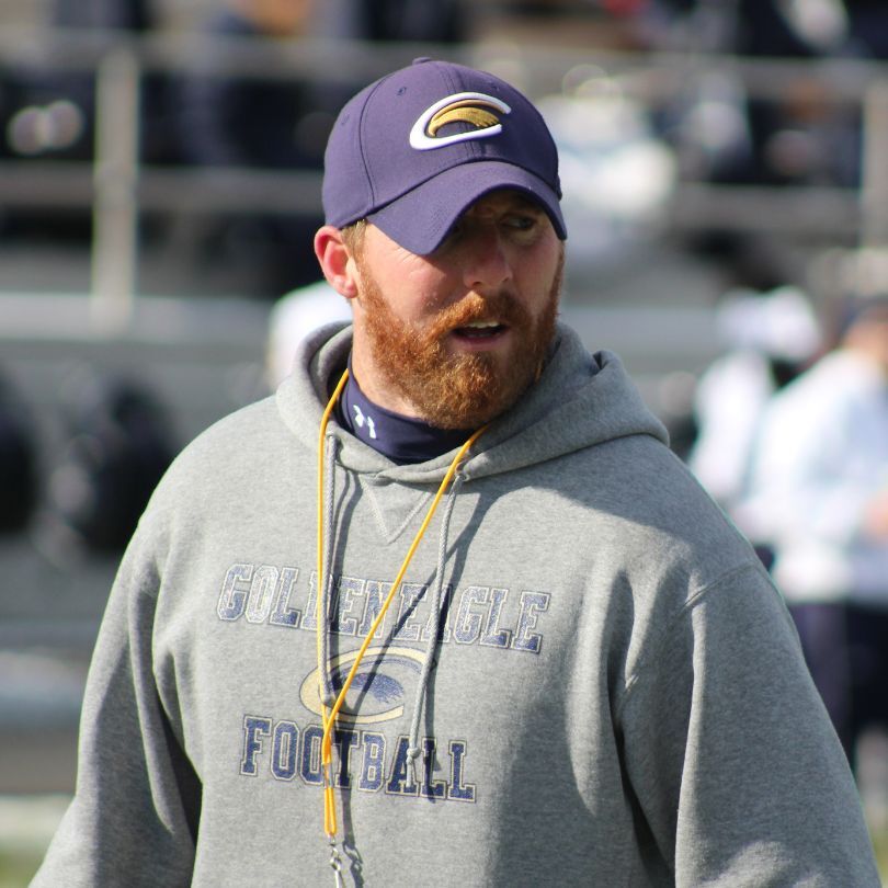 A football coaching on the sidelines of a football game, wearing a blue hat and a grey hoodie sweatshirt, watching the game with focused eyes.