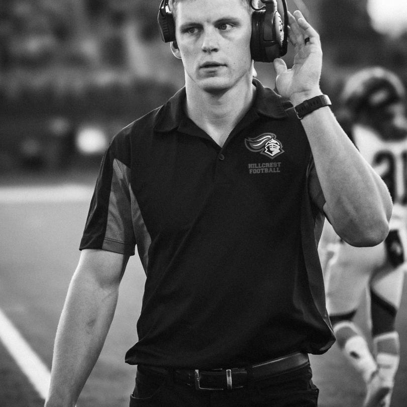 A football coaching calling plays from the sidelines and listening to communication from his other coaches via his headset. He wears a team polo.