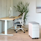 ofinto Büro-Korpus Weiss mit grauem Sitzkissen neben höhenverstellbarem Schreibtisch - minimalistisches Home-Office-Setup mit weissem ergonomischem Bürostuhl, Holzschreibtischplatte und grosser Grünpflanze, modernes Arbeitsumfeld