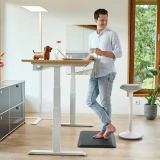ofinto Ergonomische Stehmatte Schwarz für gesundes Stehen - lächelnder Mann mit Kaffeetasse steht am weissen höhenverstellbaren Schreibtisch Elevate auf schwarzer Anti-Ermüdungsmatte in hellem Home Office mit Sitz-Stehhocker Move
