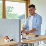 Mann im blauen Hemd arbeitet im Stehen am hellen Home Office Arbeitsplatz mit Akustik-Trennwand und Fensterblick