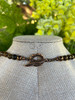 Close-up of the back clasp on the Canyon Echo necklace, featuring a bronze toggle closure with a detailed leaf motif and round Tiger’s Eye gemstone beads, displayed on a linen jewelry bust with soft natural background.