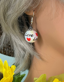 Handmade glass bead drop earrings with red hearts and ‘Love’ script design on silver-tone findings, displayed on jewelry stand.