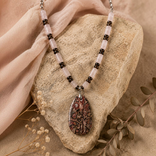 Blush of the Earth artisan necklace by Stone & Story featuring soft rose quartz, smoky quartz, and a leopard skin jasper pendant displayed on a natural stone with flowing fabric and botanical accents.