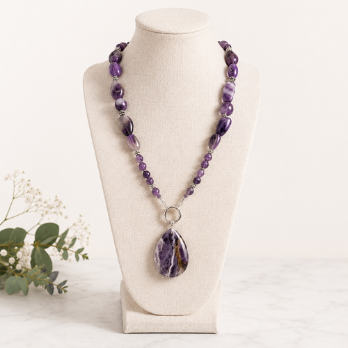 Purely Amethyst Within handcrafted gemstone necklace displayed on an ivory jewelry bust featuring genuine amethyst nuggets, polished amethyst beads, a silver toggle clasp, and a large teardrop amethyst statement pendant.