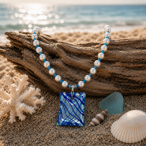 Ocean Current handcrafted necklace displayed on driftwood and beach sand, featuring luminous glass pearls, blue agate spacers, and a Murano-inspired glass pendant by Stone & Story.