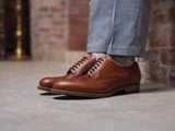 High quality natural tan full grain leather wing tip shoes three-quarter on-foot view by a brick wall - Saddleback Leather