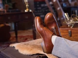 High quality natural tan full grain leather wing tip shoes upright on a fur-covered trunk with accessories - Saddleback Leather