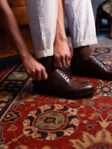 High quality dark oak full grain leather toe cap shoes being pulled on by hand, side view on oriental rug - Saddleback Leather