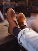 High quality tan full grain leather toe cap shoes lifestyle shot with feet resting on antique leather trunk - Saddleback Leather