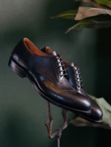 High quality black full grain leather toe cap shoes angled side profile on antique metal stand with green backdrop - Saddleback Leather