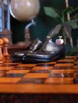 High quality black full grain leather toe cap shoes lifestyle shot on polished wooden chessboard with chess pieces - Saddleback Leather