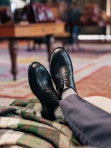 High quality black full grain leather toe cap shoes on-foot front view resting crossed on plaid wool throw - Saddleback Leather