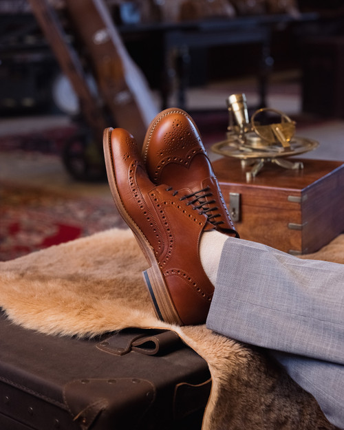 High quality natural tan full grain leather wing tip shoes upright on a fur-covered trunk with shoe care accessories - Saddleback Leather
