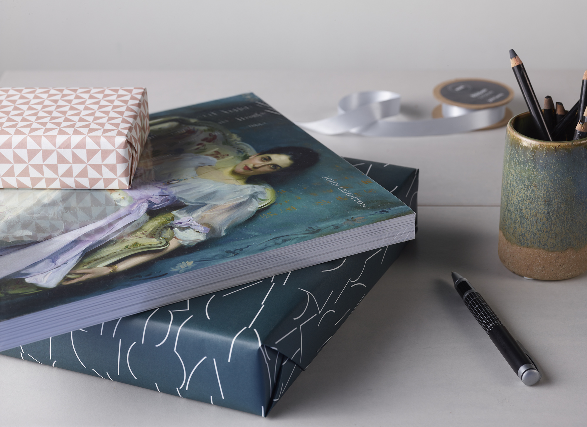 An unwrapped gallery guide book sits between two other wrapped books on a desk. Pencils and pens are nearby.