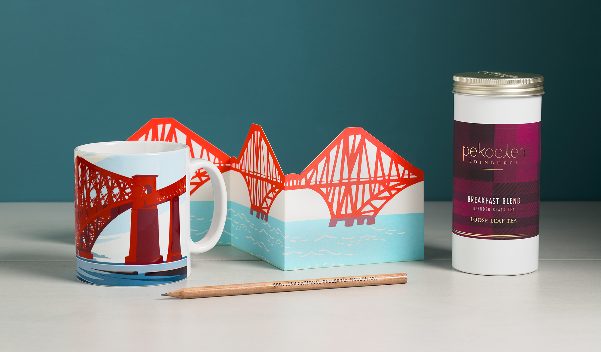 A mug and a greetings card folded in three sections sit on a white table against a petrol-blue wall. Both depict the Forth Rail Bridge in an illustrated style. Besite them, a metal tin full of breakfast tea and a wooden pencil can be seen.