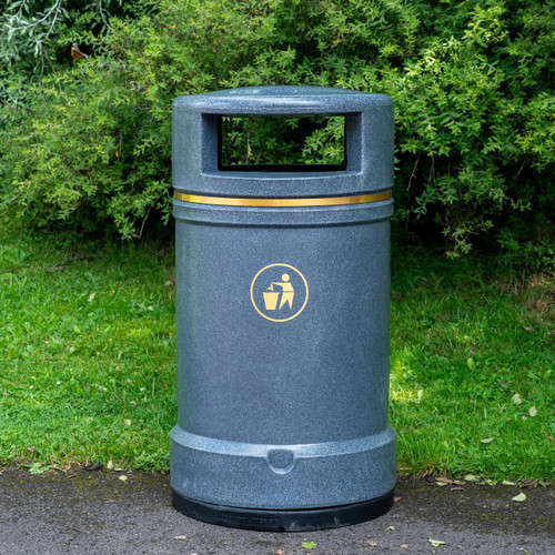 Roadware Derwent 120 Litre Outdoor Heritage Litter Bin, grey outdoor trash can with a large opening and a gold band, located on a grassy area with green bushes in the background.