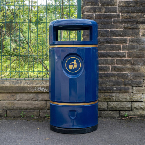 Roadware Ullswater 90 Litre Classic Heritage Litter Bin, blue public waste trash can with a recycling symbol, placed outdoors against stone wall near a green fence.